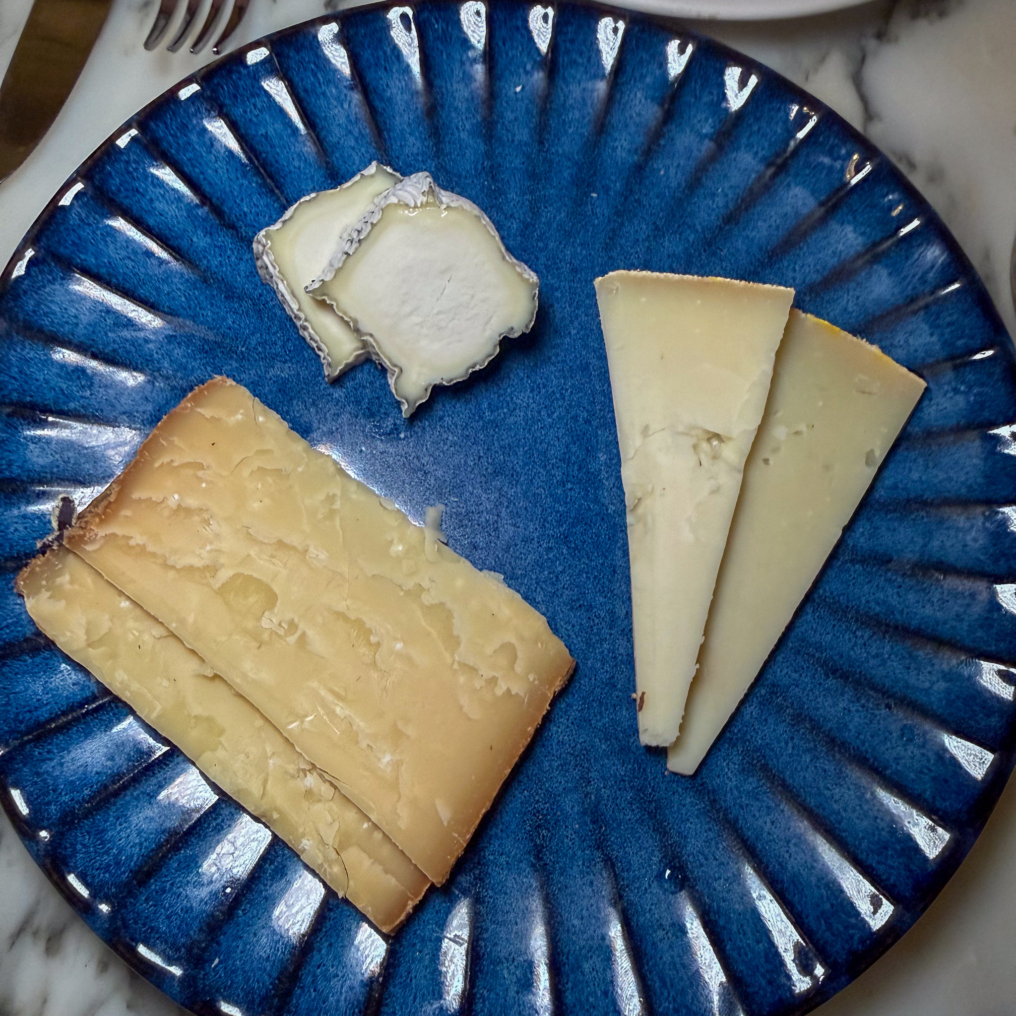 Three cheeses — a bloomy round, an aged hard wedge, and two alpine slices — on a blue plate