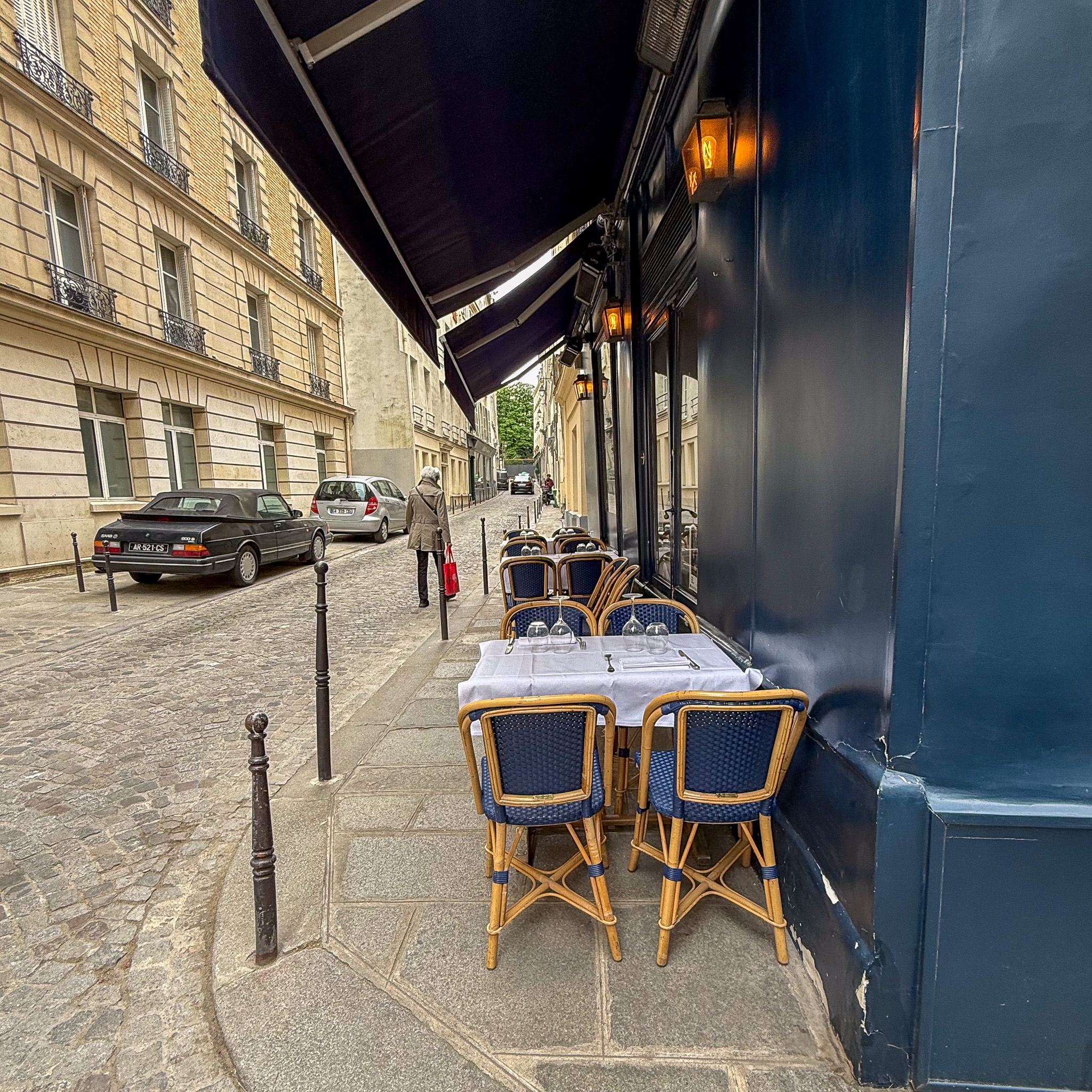 Cafe tables and wicker chairs line a narrow Parisian street with cobblestone pavement, beige buildings to the left and a dark blue storefront to the right under awnings.