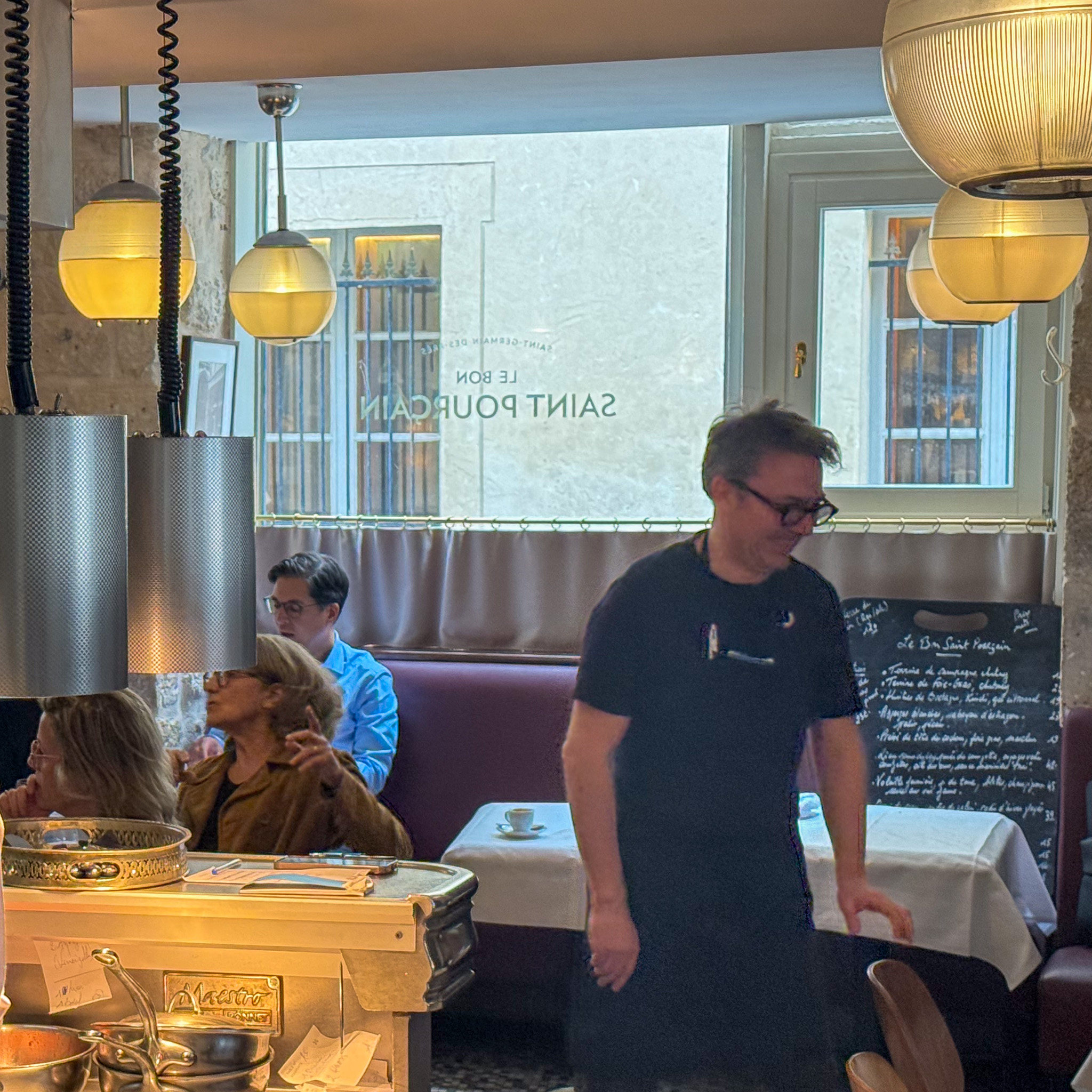 Interior of a cafe with warm yellow pendant lights, customers seated at a counter and tables, and a chalkboard menu on a wall.