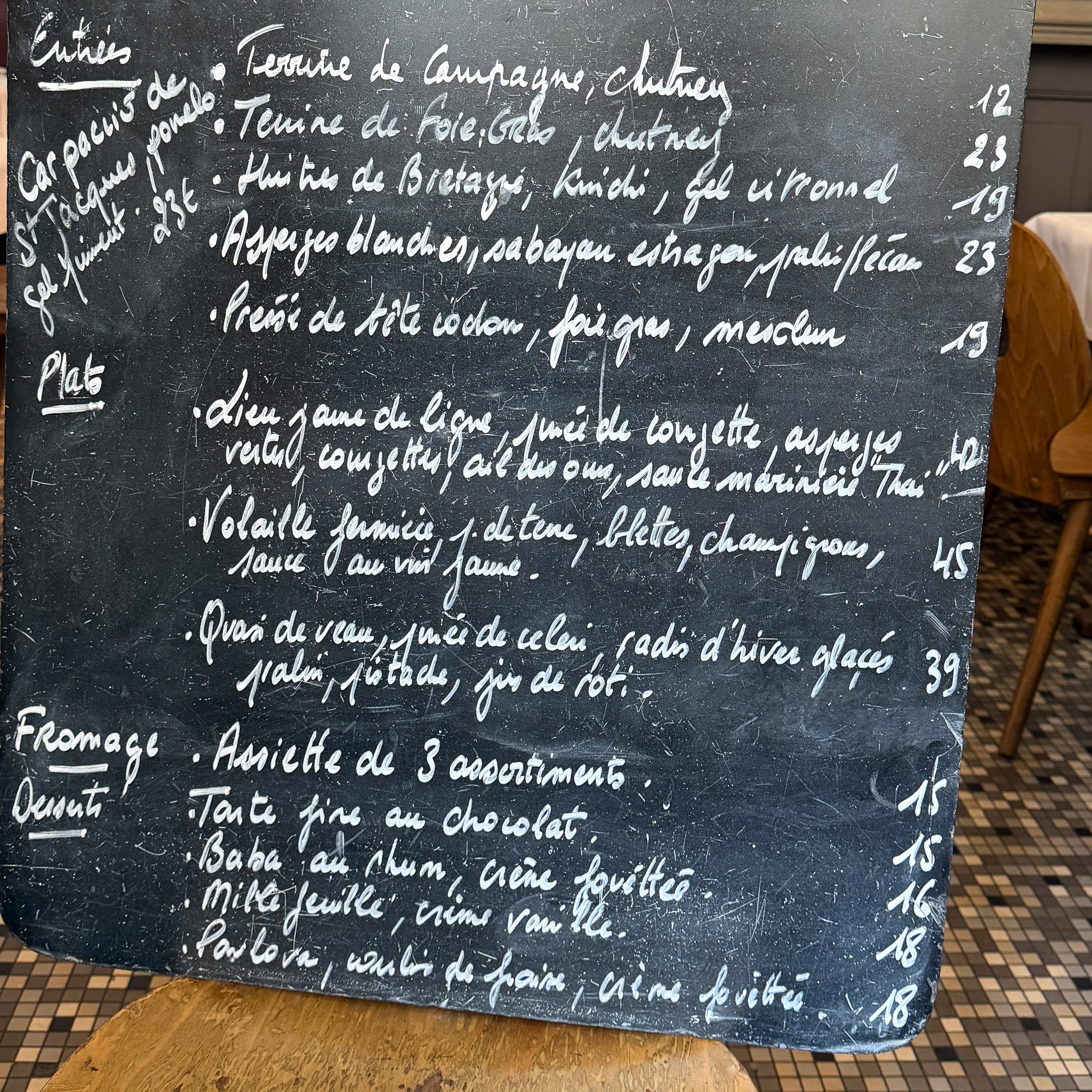 Black chalkboard menu filled with white cursive French dishes, prices listed on the right, with a wooden table and tiled floor in the background.