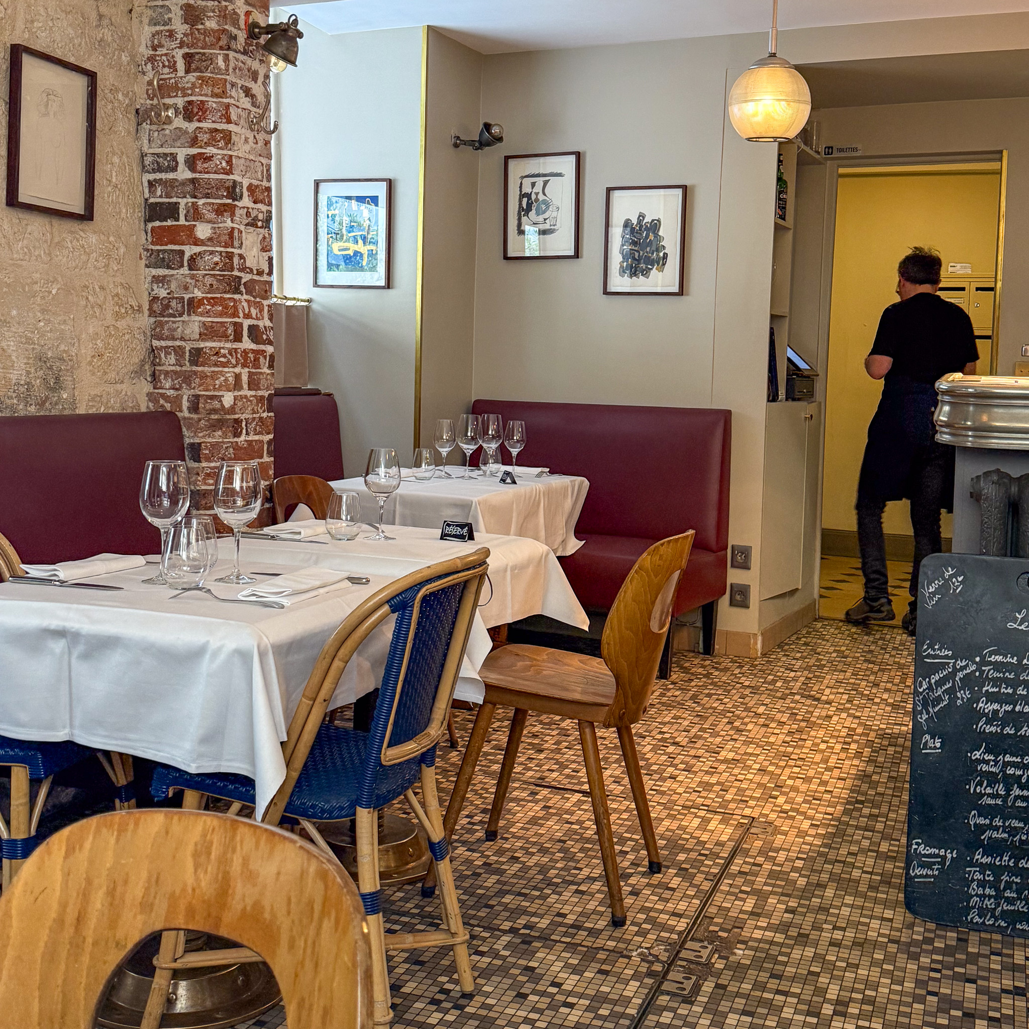 Cozy restaurant dining area with white-tablecloth tables, wine glasses, wooden chairs, a brick pillar, and framed art on beige walls.