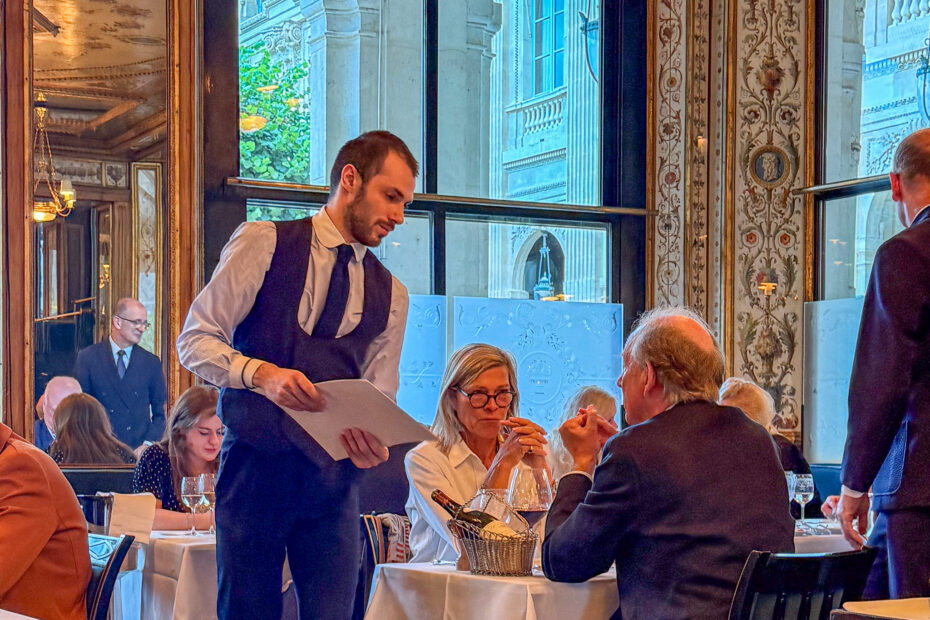 service-le-grand-vefour-restaurant-paris | PARIS BY MOUTH Waiter in vest serves a table in a formal dining room with ornate mirrored walls and chandeliers, guests seated with wine glasses.